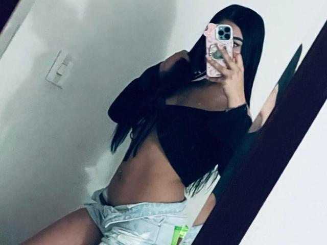 VaneVega, avec ses longs cheveux noirs, son haut noir à une épaule et son short en jean, se prend en photo devant un miroir, mettant en valeur sa silhouette fine et tonique. Ses seins sont partiellement visibles, et elle tient un téléphone avec une coque rose..