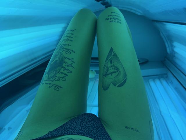 Le gambe verdi di SensualMila in un lettino abbronzante, che mostrano tatuaggi su entrambe le cosce, indossa biancheria intima a leopardati, con una luce di colore blu. Le sue cosce sono leggermente divaricate, mostrando la sua vulva..