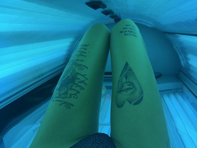 Le gambe abbronzate di SensualMila, distese su un lettino abbronzante, mostrano un tatuaggio a forma di cuore sulla gamba destra e un tatuaggio con scritte sulla gamba sinistra, con una luce UV blu che mette in risalto la sua pelle..