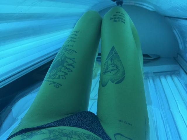 Le gambe abbronzate di SensualMila, distese su un lettino abbronzante, mostrano tatuaggi su entrambe le cosce, con il suo perizoma nero visibile, le gambe rivolte verso l'alto, sotto una luce UV blu..