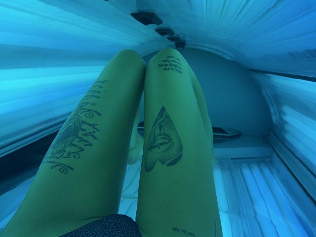 SensualMila's green legs in a tanning bed, showing tattoos on both thighs, with blue UV light highlighting her skin. Her legs are slightly parted, showing her pussy.