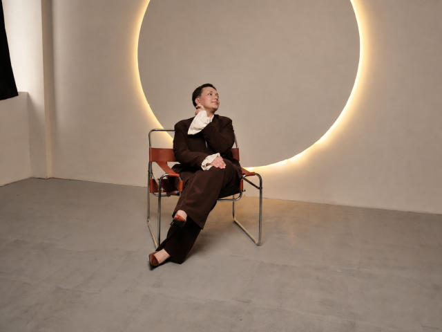 GoldenHaze with brown suit, white scarf, brown shoes, and brown pants, sitting on modern chair, hands on lap, looking up, illuminated circular wall behind, concrete floor.