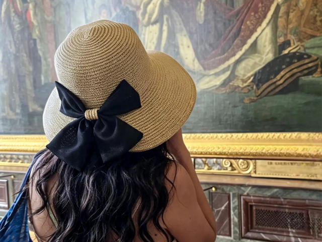 ZoeeMar, avec ses cheveux noirs ondulés, portant un chapeau de paille beige avec un nœud noir, se tient dos à la caméra, laissant apparaître ses épaules nues..