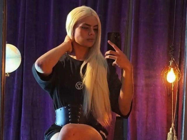 DanySky with long platinum blonde hair, black dress, and thick black corset, holding a phone, mirror selfie, purple curtain, and warm light in the background.