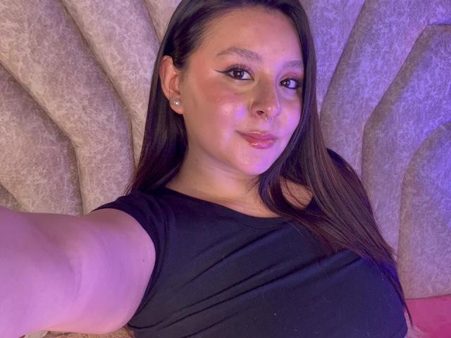 EmaJames with long brown hair, black shirt, nose ring, and diamond earrings, holding phone for selfie, showing her tits and cute smile under purple lighting.