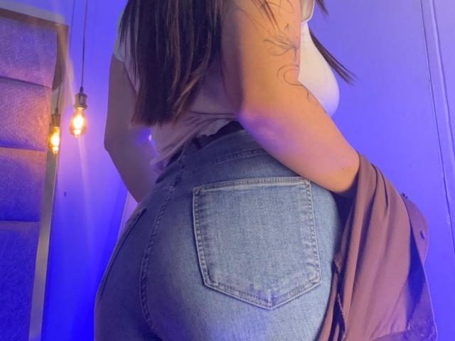 EmaJames with her butt in tight blue jeans, white top, tattoo on arm, brown hair, holding pink jacket, backlit by blue light, exposed ass pocket visible.