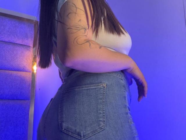 EmaJames with a white tee, denim high-waisted jeans, and a tattoo on her right arm, showing her curvy butt and hand resting on her hip. Blue lighting.