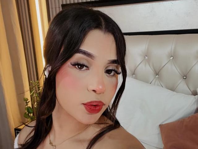 LunaVali, con i capelli scuri, il rossetto rosso e il trucco pesante sugli occhi, fa la smorfia, mostrando le sue labbra piene e rosa. Il suo piccolo sedere rotondo è leggermente visibile sul letto bianco..