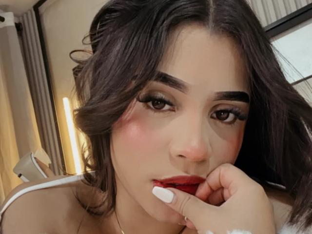 LunaVali a les cheveux noirs et ondulés, des sourcils épais, du rouge à lèvres et des ongles blancs qui touchent légèrement sa bouche entrouverte. Sa main repose sur sa joue dans une pose séduisante..