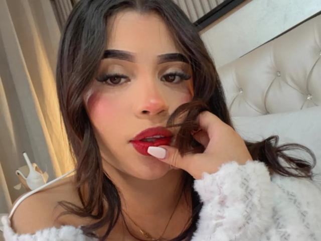 LunaVali, con i capelli scuri, il rossetto rosso e il maglione bianco e soffice, tiene in mano una ciocca di capelli, mostrando lo smalto rosa alle unghie e guardando la fotocamera con un leggero sorriso..