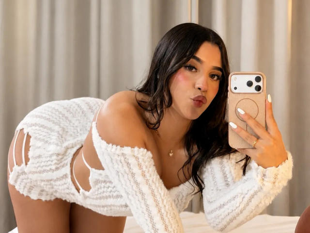 LunaVali, avec ses longs cheveux foncés, portant une mini-robe blanche en tricot, avec une épaule dénudée et des découpes sur les côtés, est penchée en avant, prenant un selfie avec un iPhone rose, montrant ses fesses..