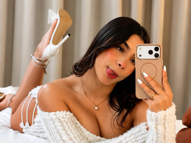 LunaVali, con i capelli scuri, la lingua fuori, che tiene un telefono, indossa una maglia bianca a spalle scoperte, mostrando il seno, scarpe con il tacco bianco e braccialetti, sdraiata su un letto..