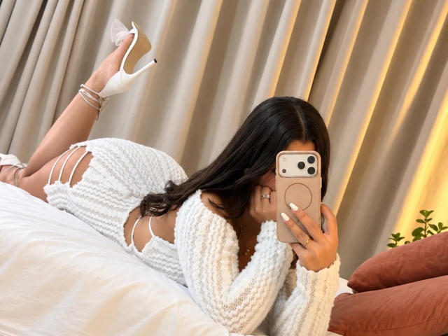 LunaVali, allongée sur un lit, portant une robe blanche en maille côtelée avec des découpes sur les côtés, et des talons hauts blancs, prend un selfie avec son téléphone, en cachant son visage..
