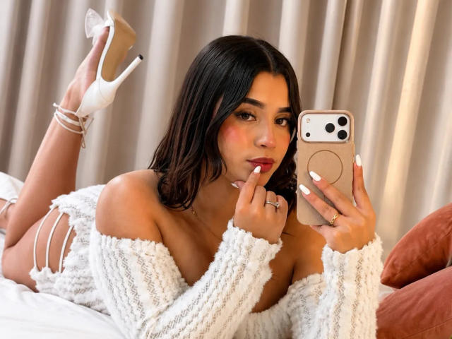 LunaVali with medium tits, lying on a bed, wearing a white knit off-shoulder sweater and white high heels, taking a selfie with a beige phone.
