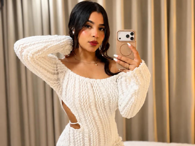 LunaVali, avec ses cheveux foncés, portant un pull en tricot blanc texturé, à épaules dénudées, prend un selfie avec un téléphone beige, montrant des découpes sur les côtés, tout en remontant ses cheveux et en regardant l'appareil photo..