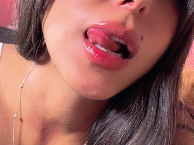 Os lábios rosados e brilhantes de SarahPrincess estão ligeiramente entreabertos, revelando uma pequena marca de beleza preta no lado esquerdo. Sua língua está parcialmente para fora, e seu cabelo castanho escuro é visível..