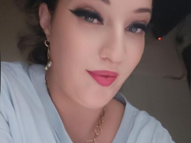 VanessaSquirtt with red lipstick, black eyeliner, and blue shirt, wearing gold earrings and necklace, has light skin, and brown hair, smiling softly at the camera.