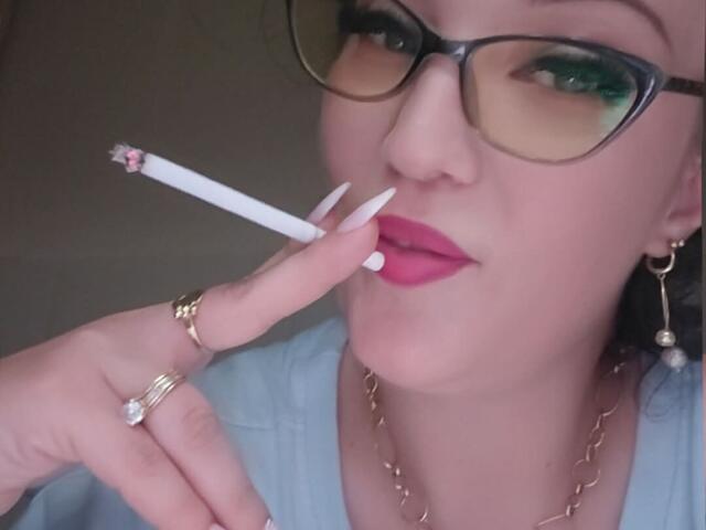 VanessaSquirtt with glasses, pink lipstick, white nails, holding a cigarette, gold rings, necklace, and earrings, looking at the camera with a slight smile.
