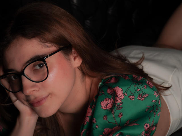 LiaSwann con gli occhiali, una maglietta verde con fiori, pantaloni bianchi, sdraiata su pelle nera, viso leggermente arrossato, capelli castani, sorriso appena accennato, mano sotto il mento, illuminazione soffusa..