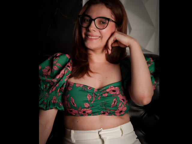 LiaSwann, avec des lunettes, un haut court vert à motifs floraux qui révèle ses seins, un pantalon blanc, souriante, cheveux bruns, une main sur la joue, fond noir, et une chaise en cuir noir..
