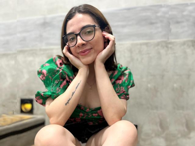 LiaSwann, avec des lunettes, un haut vert à motifs floraux, les mains sur les joues, souriante, un tatouage sur l'avant-bras droit, cheveux bruns, lunettes à monture noire, assise, fond gris et blanc..