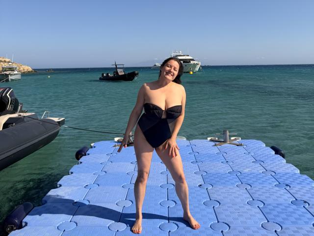 BadAngel avec de grosses seins et un maillot de bain une pièce noir, debout sur un tapis bleu flottant dans une eau claire, souriante, les jambes écartées, des bateaux en arrière-plan, une journée ensoleillée..