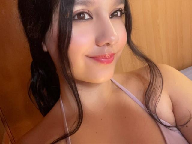 Jade Palacios, avec ses cheveux noirs, sa peau claire et son rouge à lèvres rose, souriante, montrant de petites seins dans un haut à encolure plongeante rose, fond en bois, autoportrait..