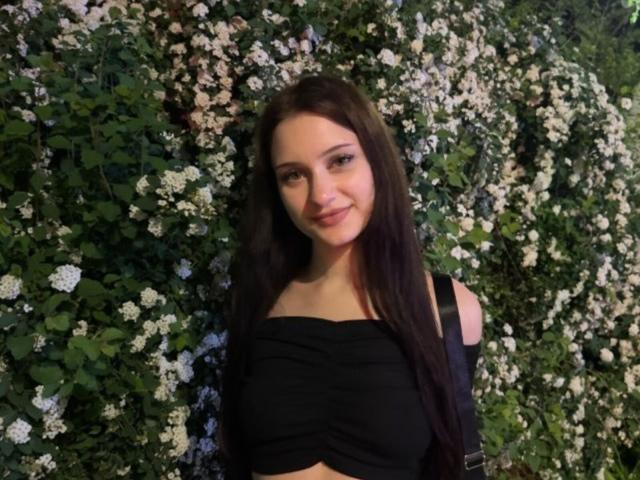EleanorEvanchyk with long brown hair, smiling, wearing a black off-shoulder crop top, standing in front of a green, white-flowered wall. Boobs visible, cute face, natural look.
