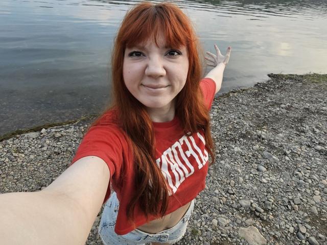 HalleyBabiracki with red hair, red crop top, and blue jeans, smiling and taking a selfie by a rocky water edge, right arm extended, left arm pointing.