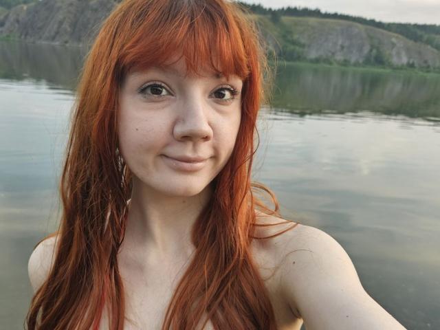 HalleyBabiracki has red hair, freckles, and is topless with small tits, a nose piercing, and a slight smile, standing by a calm lake with a rocky hill in background.