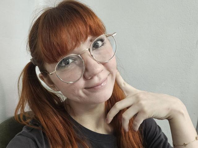 HalleyBabiracki with red hair in pigtails, wearing round glasses, smiling slightly, touching her neck with a hand, wearing a grey shirt, and a subtle smile on her face.