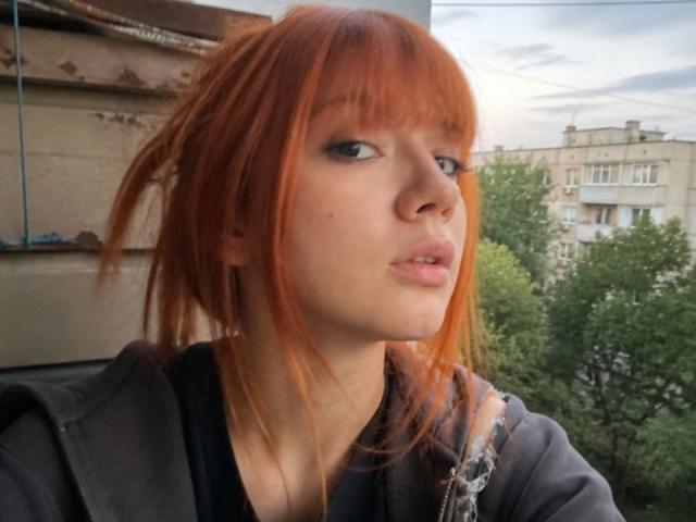 HalleyBabiracki has red hair, blue eyes, and is pouting with slightly parted lips, wearing a black shirt, looking to the side. She's outside with green trees in the background.