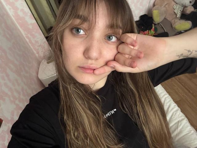 MichikoVivona has long brown hair, blue eyes, and a black shirt. She's touching her left cheek with her right hand, showing a small tattoo on her wrist.