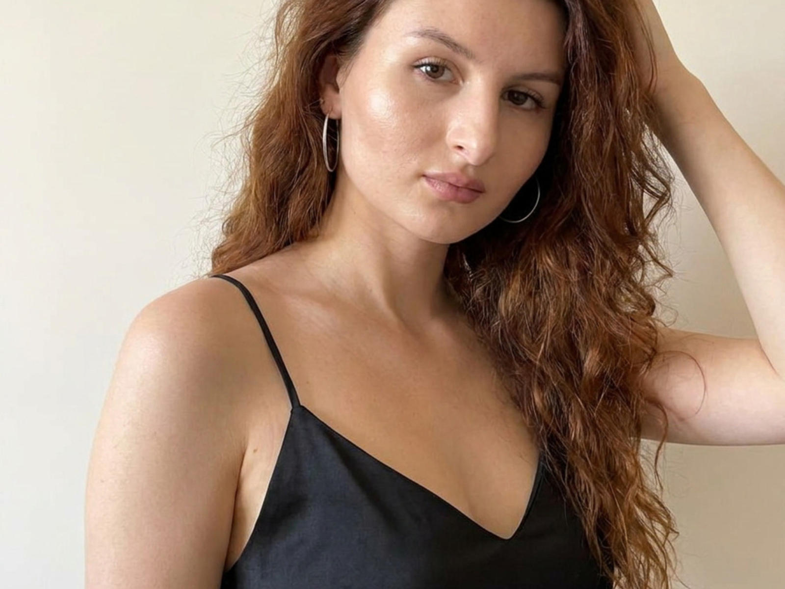Genevieve Groshans, com cabelos castanhos ondulados, vestindo uma blusa preta de alças finas, tem seios pequenos, brincos de argola e um sorriso discreto, em um fundo simples..