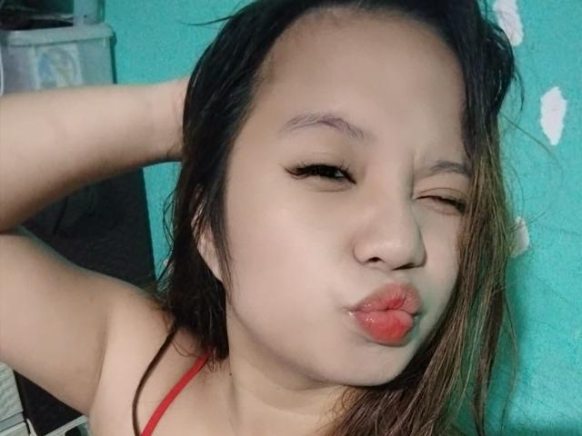 YannaDirtyOne with one eye closed, puckering her red lipstick-covered lips, has her hand in her dark, wet, shoulder-length hair, wearing a red strap top.