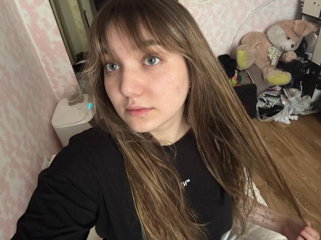 MichikoVivona with long brown hair, blue eyes, and a serious expression, wearing a black shirt, in a bedroom with stuffed animals and clothes on the floor.