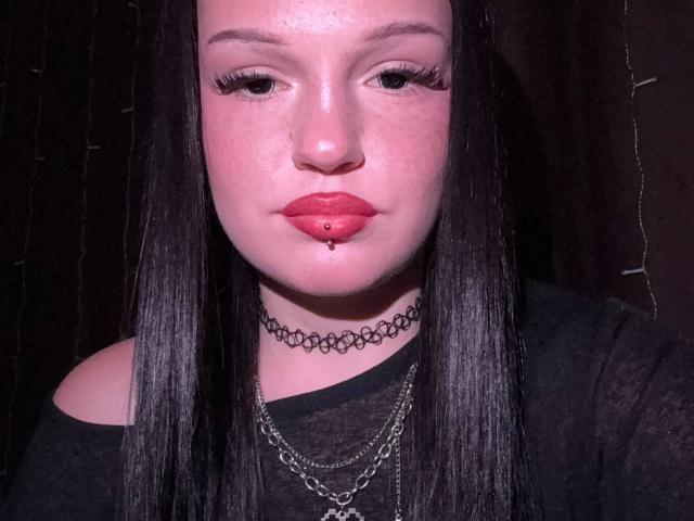 HolliKendig has pale skin, black straight hair, red lipstick, a black choker, and a chain necklace. She's looking down with a slight pout, showing a pierced lower lip.