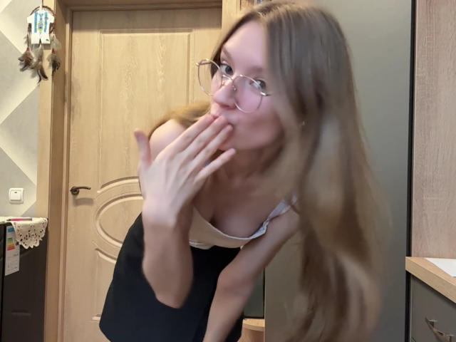 ColeneBeauharnois with long blonde hair, glasses, and a white tank top, covering her small tits with her hand, smiling, in a blurry kitchen scene.
