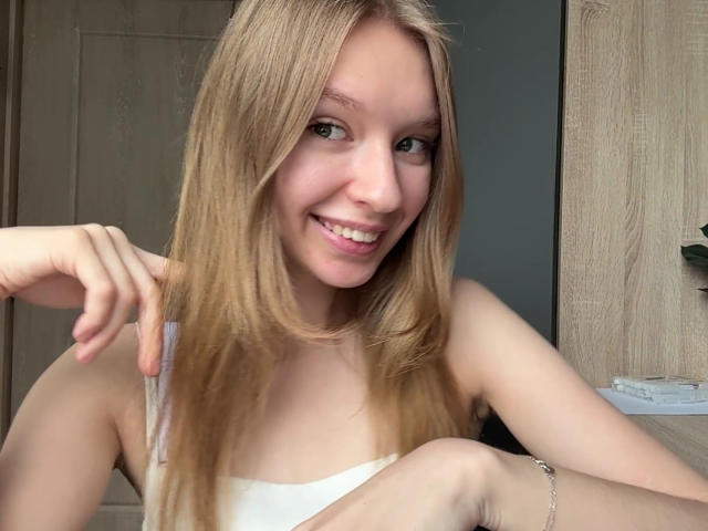 ColeneBeauharnois with long blonde hair, smiling, wearing a white tank top, holding her hair with one hand, showing her ass slightly, sitting, light skin, green eyes, and a bracelet.