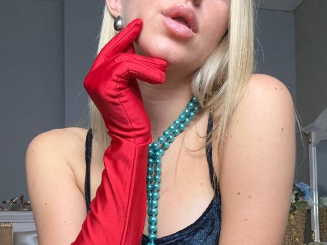 BalletSwan with blonde hair, wearing red satin gloves, teal bead necklace, black tank top, and black earrings, touching her chin with red-gloved hand, showing her pouty lips.
