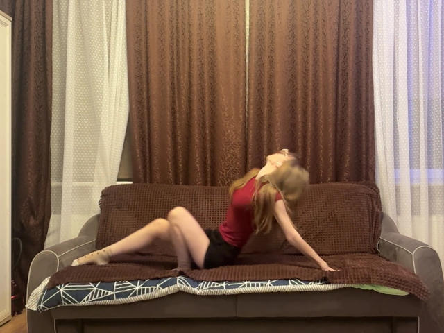 ColeneBeauharnois with red top and black shorts, arching back, hands on brown sofa, legs spread, blonde hair, eyes closed, brown curtains behind, white sheer curtains on sides.