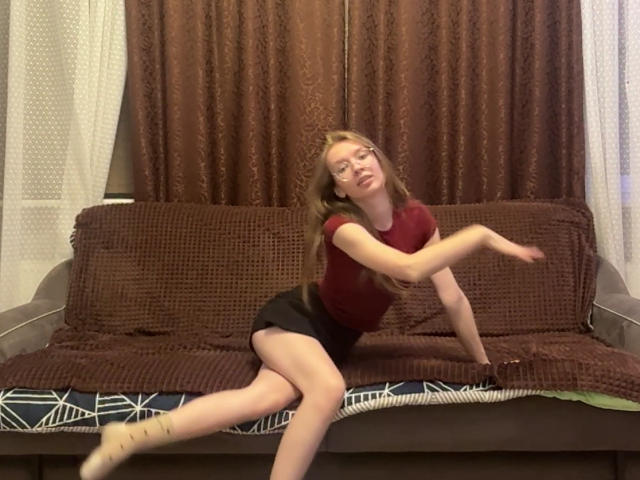 ColeneBeauharnois, with glasses and long brown hair, is on a brown couch, wearing a red shirt and black shorts, in a dynamic, almost dancing, pose with her arm out.