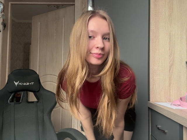 ColeneBeauharnois with long blonde hair, red shirt, leaning forward, showing her small tits, smiling, in front of a black X-Force gaming chair.