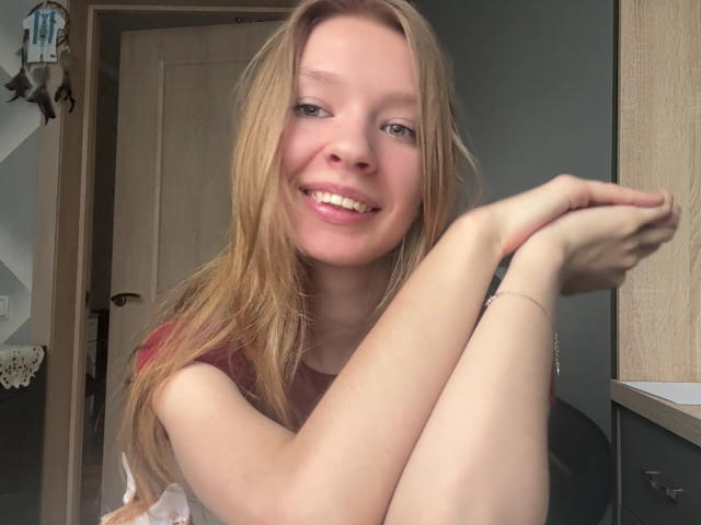 ColeneBeauharnois has long, messy blonde hair, a cute smile, and is sitting with her arms crossed, showing off her small tits and a slender, pale body.