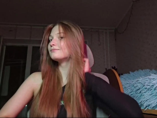 JudieSlowey, avec de longs cheveux bruns et lisses, portant un haut noir, avec un léger sourire, tenant ses cheveux relevés, assise sur une chaise avec un coussin bleu duveteux..