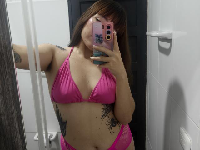 ValeriHairy69 with pink bikini top and matching pink mesh bottom, holding phone with pink case, showing tattoos, and covering her mouth with phone. Her tits are small.