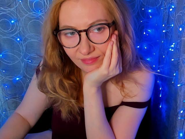 PaulinaSunny with glasses, blonde hair, and fair skin, resting her cheek on her hand, wearing a black top, blue fairy lights in the background, smiling softly.