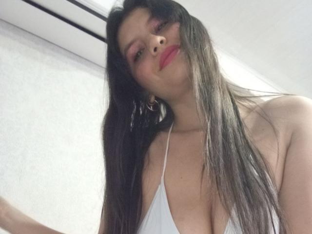 Madisson Fontaine, avec ses longs cheveux noirs, son rouge à lèvres rouge et son haut blanc à encolure dégagée qui révèle ses seins. Elle regarde vers le bas avec un léger sourire. Un climatiseur se trouve au-dessus d'elle..