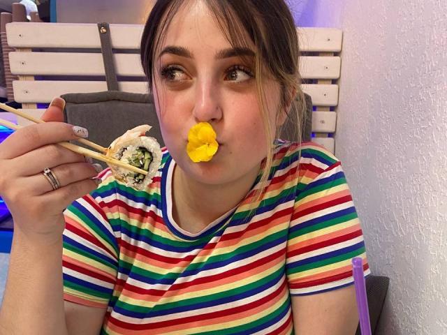 AddieLoretta, avec ses lèvres jaunes et gonflées, mange un pain avec des baguettes, porte une chemise rayée de toutes les couleurs de l'arc-en-ciel, a les cheveux bruns attachés en un chignon désordonné et porte une bague à la main gauche..
