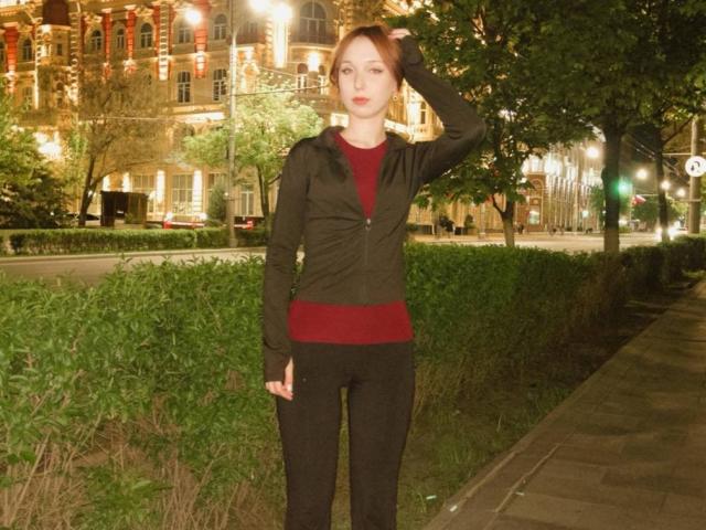 EstelaYake stands with hand in red shirt, black pants, and green vest, against night-lit urban park. She's got pale skin, red hair, and a serious face.